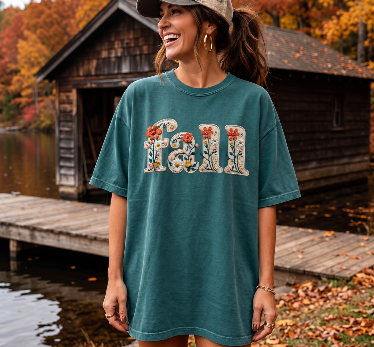 Fall Bloom Tee in Blue Spruce with cream floral fall lettering on a model beside an autumn cabin.